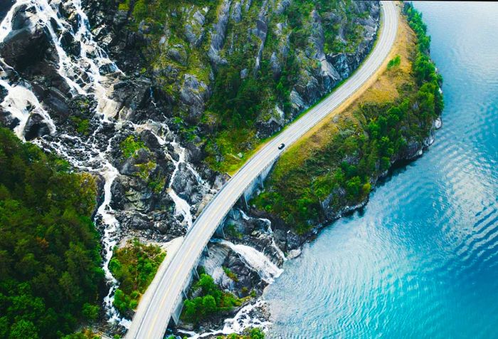 A car navigates a coastal road, crossing a hillside waterfall that cascades into the ocean.