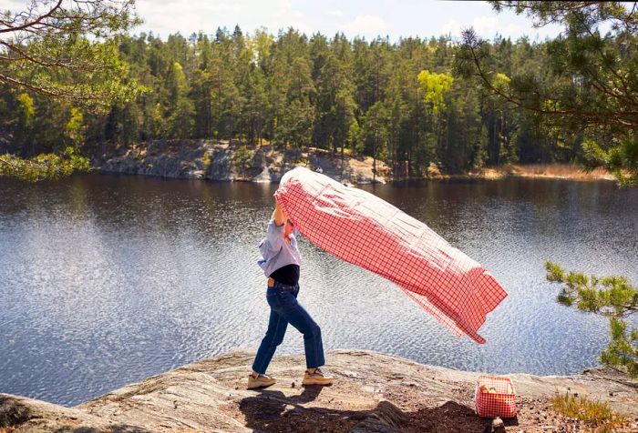 A woman with a pink picnic blanket sitting on a cliff by the lake, surrounded by tall green trees.