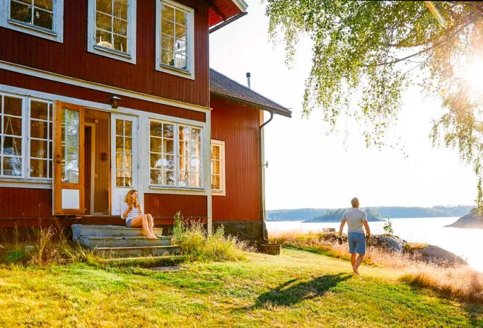 A woman perched on a staircase outside her home, observing a man walking towards a lake.