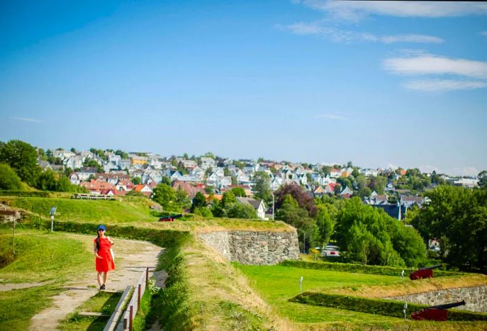 A woman in a vibrant red dress stands atop a fortress, gazing over a cluster of buildings in the distance.