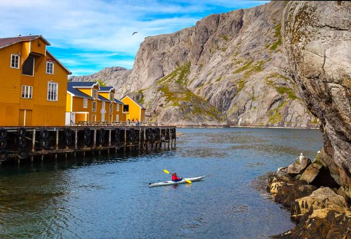 A man paddles a kayak in a serene fjord next to a quaint yellow fishing village, surrounded by towering rocky mountains.