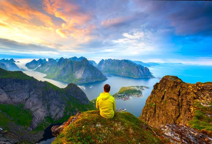 A man perches on a rocky peak, enjoying a breathtaking view of steep islands rising from the sea beneath a picturesque cloudy sky.