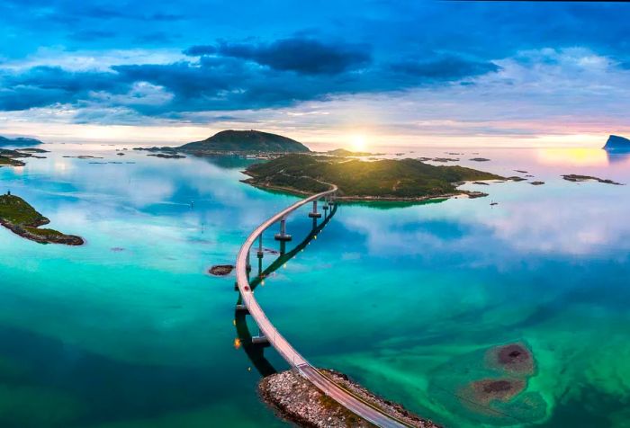 The expansive Tromsø bridge stretches gracefully over the turquoise strait, bathed in sunlight against a backdrop of cloudy skies.