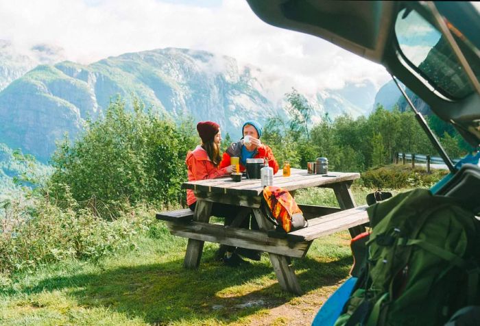 A couple dressed in winter attire enjoying breakfast at a picnic table surrounded by mountains.