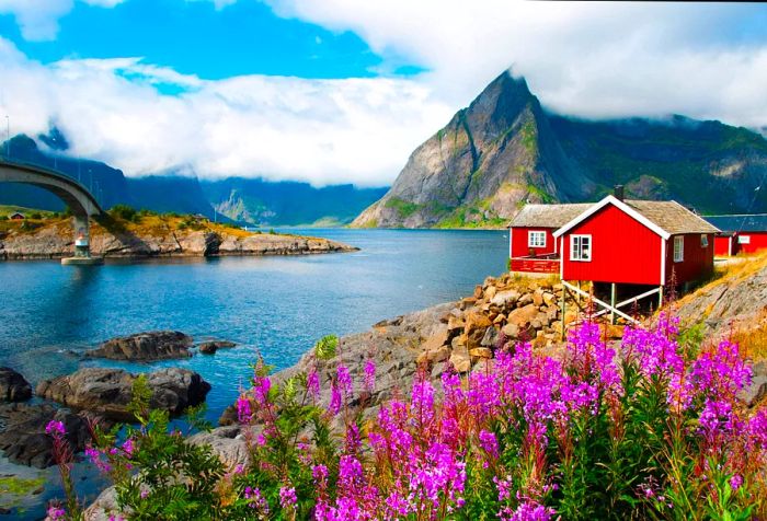 A cluster of red wooden houses perched beside a river flowing through steep mountains, connected by a charming bridge.