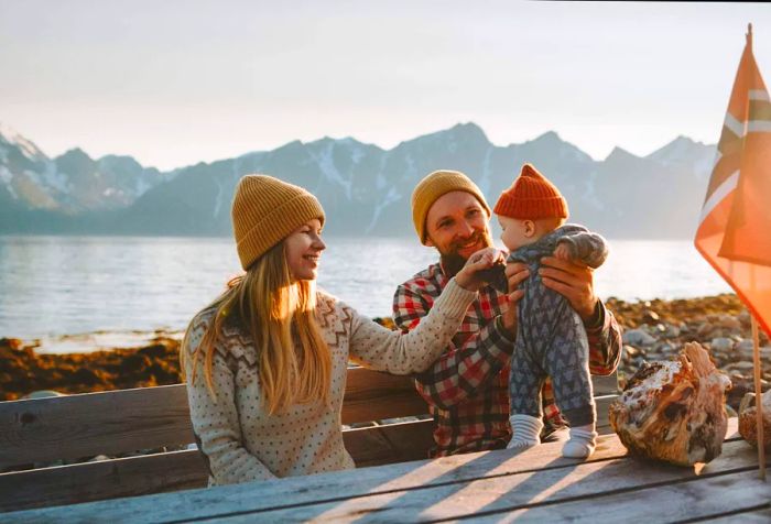 A family of three bundled in warm clothing sits at a table, with the father cradling the baby in his arms.