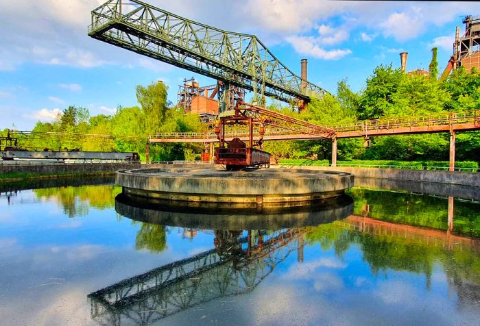 The ore loading bridge, affectionately known as 'The Crocodile,' reflects in the clarifier at Duisburg Landscape Park (Landschaftspark Duisburg-Nord). The blast furnace complex, constructed in 1902, suffered significant damage during World War II but was reconstructed in the 1950s. By 1985, the blast furnaces became unprofitable, leading to the complex's closure. From 1991 to 2002, the area was transformed into a public park.