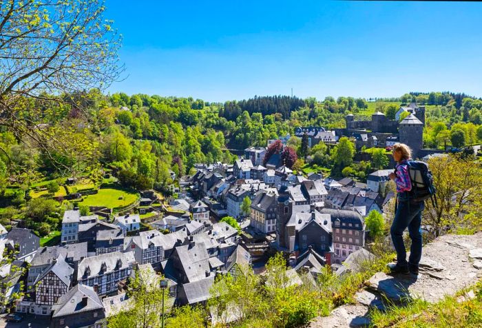 A hiker navigates a cliffside path with a view of a village nestled among thick forests.