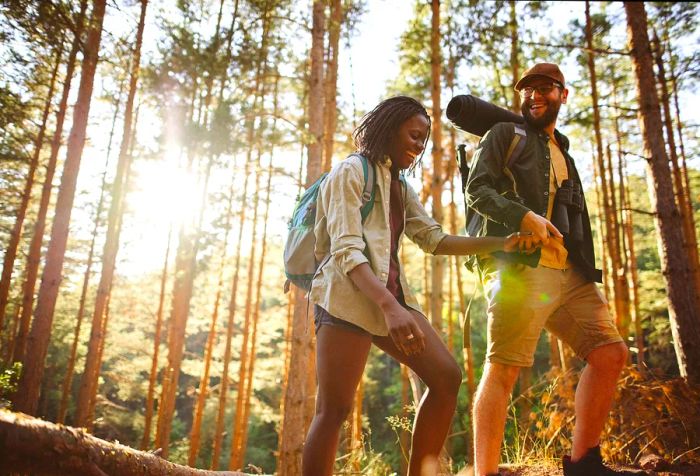 A joyful couple walks hand in hand through the woods, carrying camping bags.