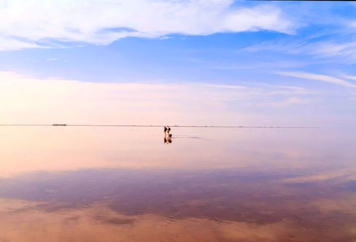 A couple and their dog stroll across a mudflat that reflects the sky above.