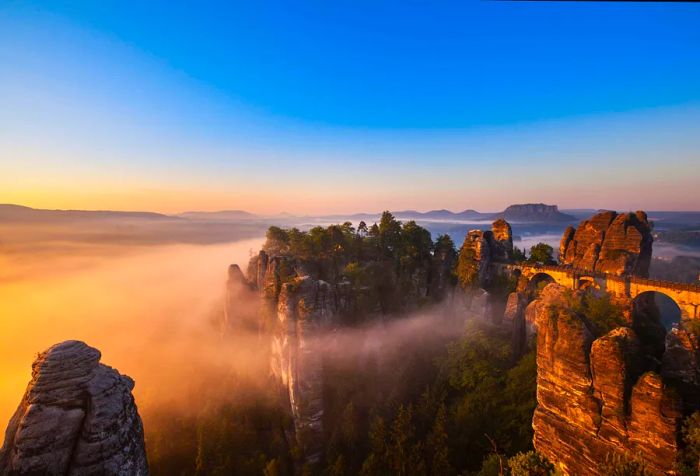 A bridge spanning a rugged mountain ridge leads to mist-covered, eroded rock formations.