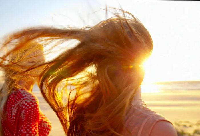 A woman's golden hair dances in the breeze as she stands beside another woman on the beach.