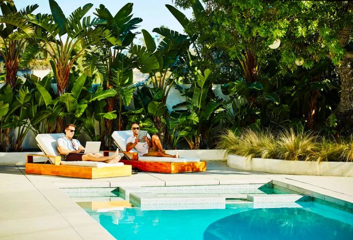 A gay couple lounging by the pool, using their laptop and tablet.