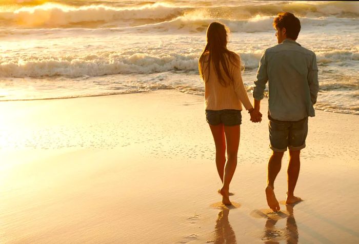 A couple is seen from behind, holding hands as they stroll along the beach with large white waves crashing nearby.