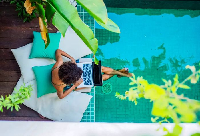 A woman perched on large cushions by the pool, her feet playfully splashing in the water as she works on her laptop.