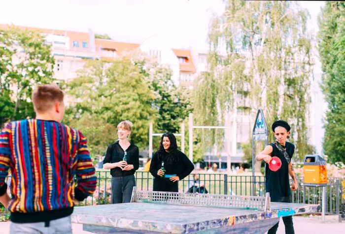 Two women enjoying drinks while watching a couple of guys play ping-pong.