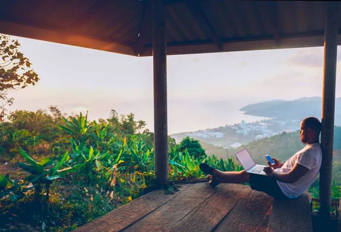 A male traveler seated on a wooden shed, working on his laptop and phone, overlooking a village and the ocean below.