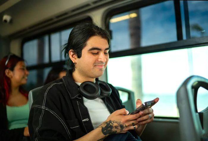 A young man on a bus, headphones draped around his neck, completely engrossed in his smartphone.