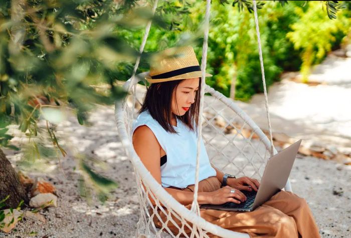 A woman working on her laptop, comfortably seated on a swing chair suspended from a tree by the beach.