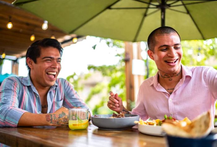 Two male partners share a laugh while seated at a table filled with food and drinks.