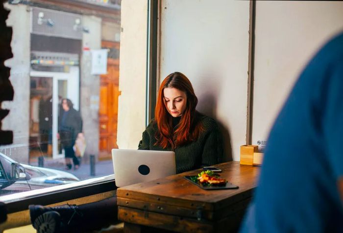 A long-haired woman works comfortably on her laptop by a large glass window in a cozy café.