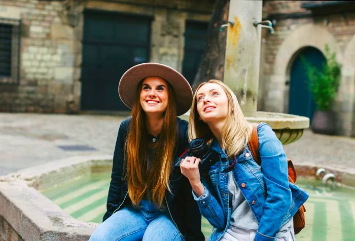 Two young women sit by a fountain, looking up, one sporting a beautiful hat while both are dressed in cozy jackets.