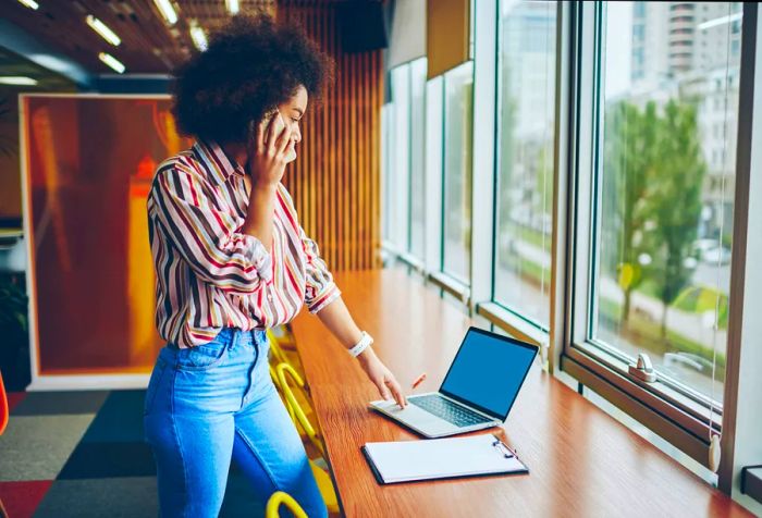 A woman with stunning afro hair stands, chatting on her phone while glancing at her laptop on the table.