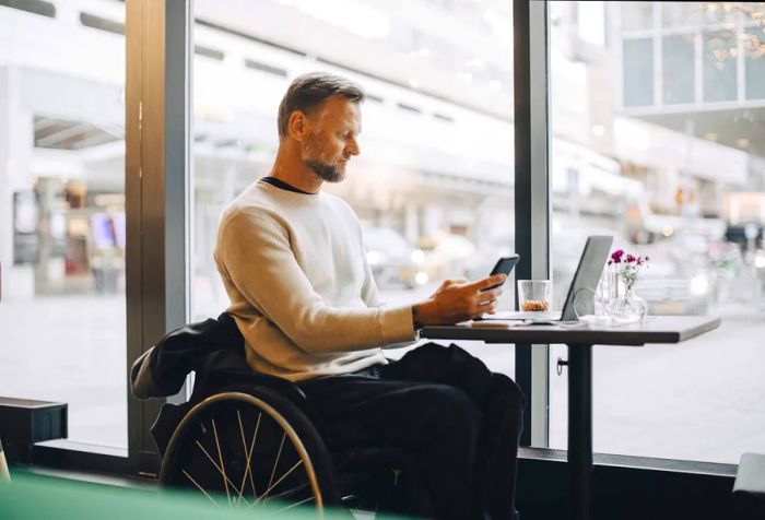 A man in a wheelchair checks his mobile phone while his laptop sits on a table in a restaurant.