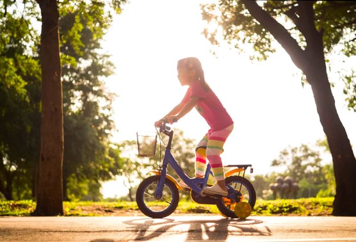 A girl rides her small bike along a sun-drenched asphalt road bordered by tall trees.