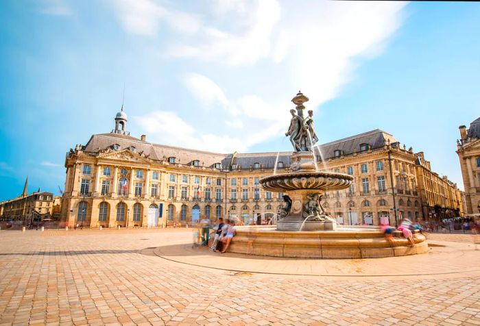 The Three Graces Fountain stands prominently in La Bourse square, with the Palais de la Bourse visible in the background.