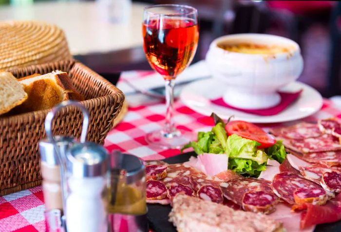 On a table adorned with a checkered cloth, there were cured meats, a glass of wine, bread, and soup.