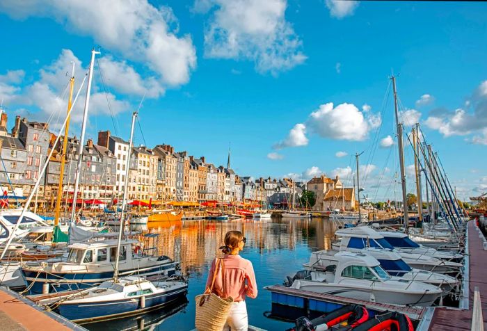 A woman holding a rattan bag stands on a pier, gazing out at a marina filled with docked boats.