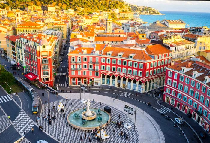 A round fountain featuring a statue sits in an open square, surrounded by red-hued buildings and a distant glimpse of the sea.