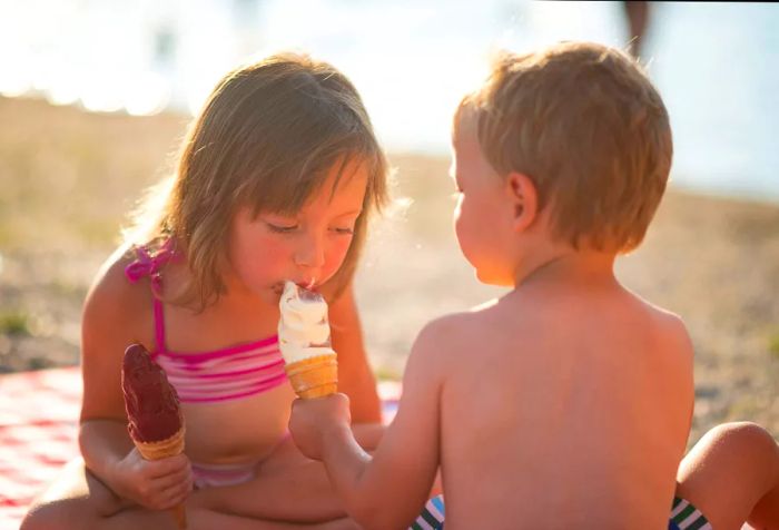 Siblings share a joyful moment on the beach while enjoying ice cream cones, with one playfully tasting the other's treat as they happily share.