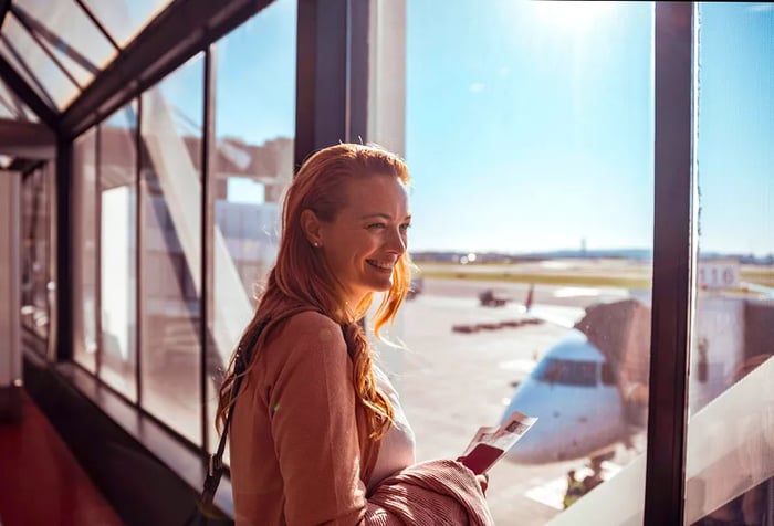 A woman beams while standing by the large glass windows of an airport terminal, holding her ticket.