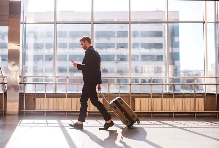 A male traveler checks his smartphone while strolling through the airport with his luggage.