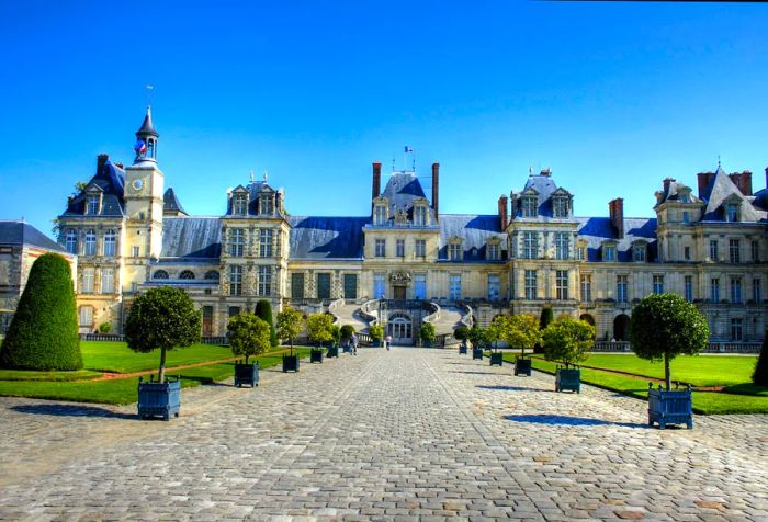 A cobblestone path adorned with potted plants and lush green lawns leads to the Palace of Fontainebleau, a medieval manor featuring an outdoor double staircase, numerous windows, and blue-tiled roofs.