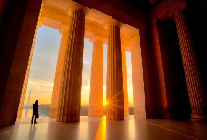 Sunlight streams through the tall Doric-style columns, illuminating a lone figure standing below.