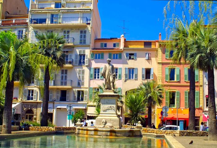 A statue perched above a lion over a pond, encircled by palm trees and a row of vibrant buildings.