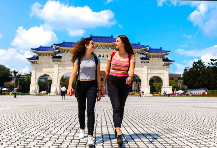 Two young women stroll across a paved plaza located in front of a traditional Chinese-style gateway.