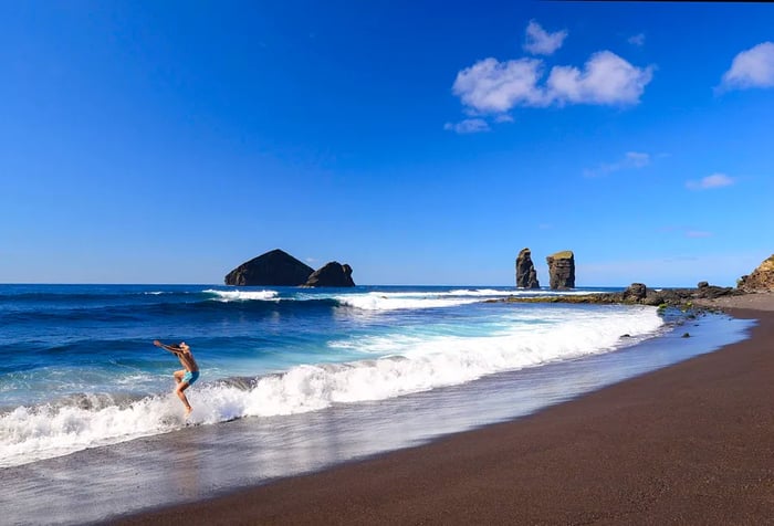 An individual leaping over ocean waves at a striking black sand beach.