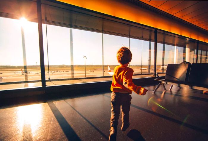 A young child in an orange sweater dashes through an airport lounge filled with large windows.