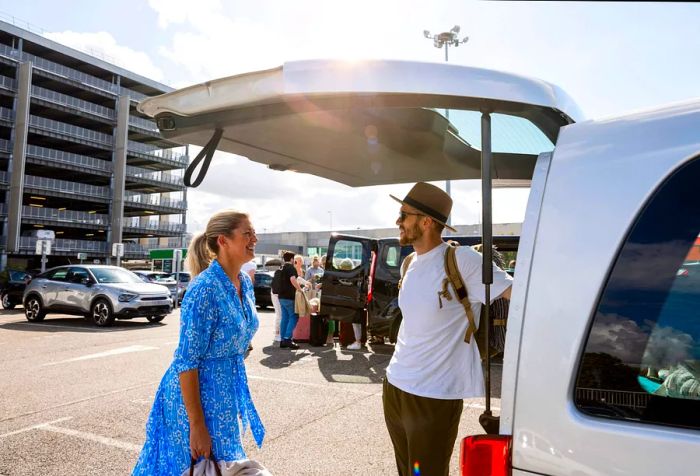 Two people are engaged in an animated discussion at the back of a white van parked in a large parking area.