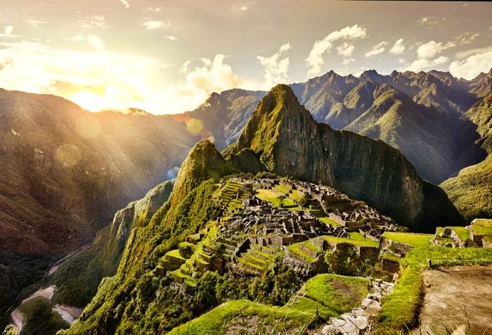 The brilliant sun shines through the open sky, illuminating the mountains that surround Machu Picchu.