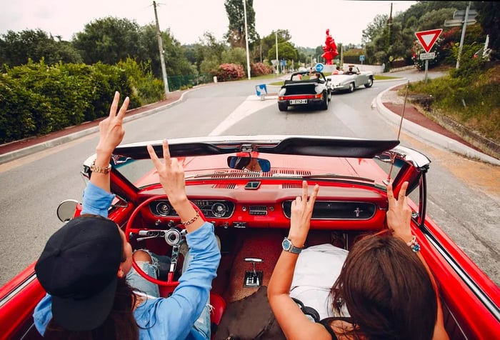 Two women in a red convertible joyfully raising their hands and flashing peace signs.