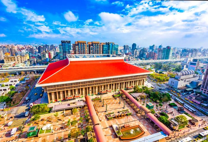 A striking building with a red roof sits beside a park featuring covered walkways, all set against the backdrop of a bustling city skyline.