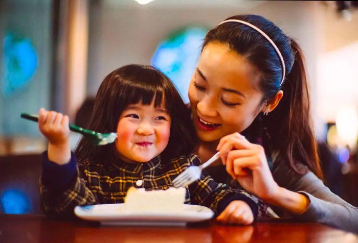 A mother and her young daughter enjoy a slice of cheesecake together.