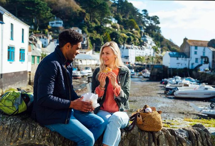 A couple enjoys a meal and conversation while perched on a rock wall, overlooking white houses nestled on the mountainside in the distance.