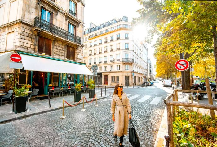 A fashionable woman strides confidently across the street, framed by the charming facade of a restaurant, while a picturesque tree-lined avenue adds to the ambiance.