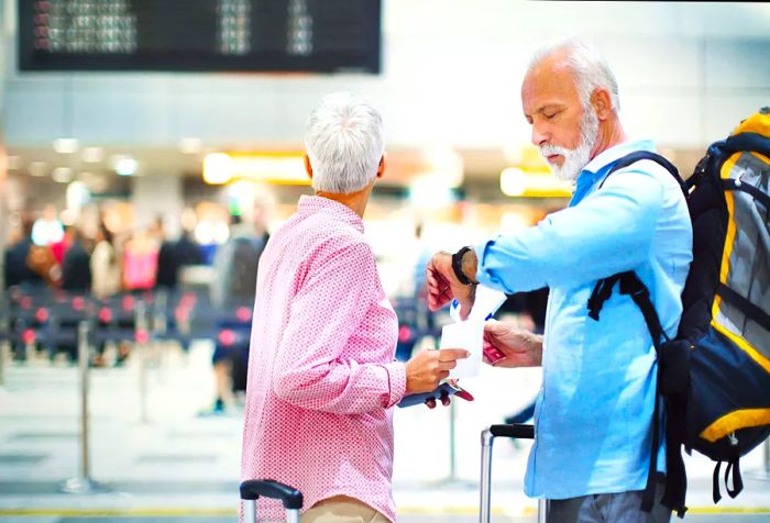 A close-up view of an elderly couple at the airport, waiting for their delayed flight. Both hold their passports and tickets, with the woman glancing at the arrival and departure board. (Note: the tickets are generic and do not contain any data.)